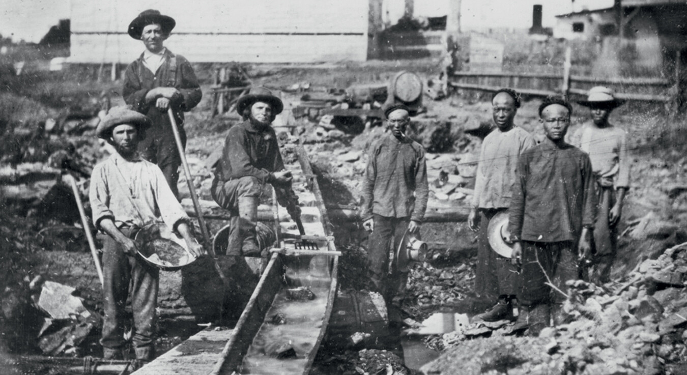 A photo of Chinese immigrants and white settlers mining for gold in the Auburn Ravine of California in 1856. Water runs through a sluice in the middle of the photo, so the miners can pan for gold in it.