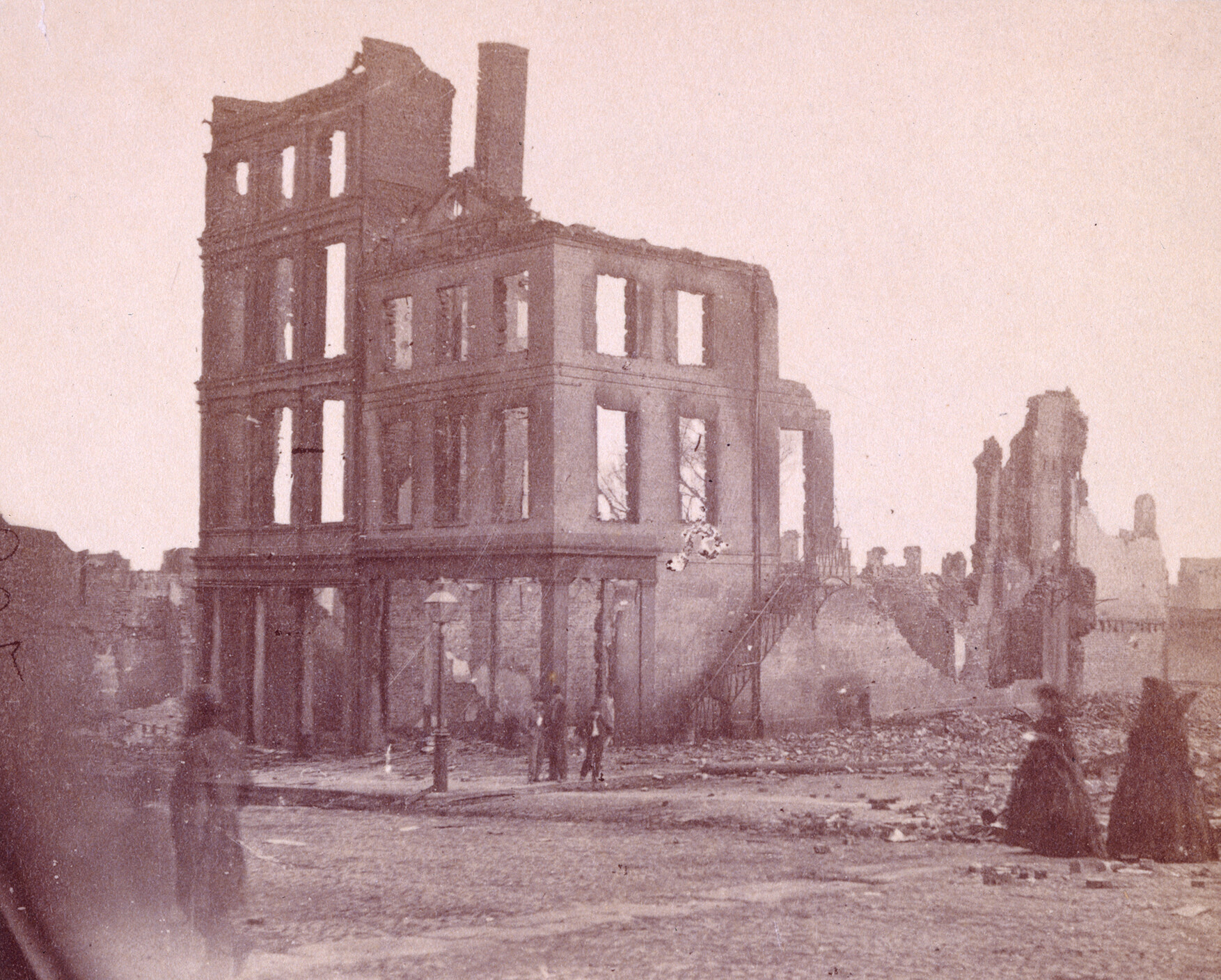 A photo shows the ruins of a building in Richmond, Virginia in April 1865. Women in mourning attire walk among the shambles while a few people gather near the rubble.