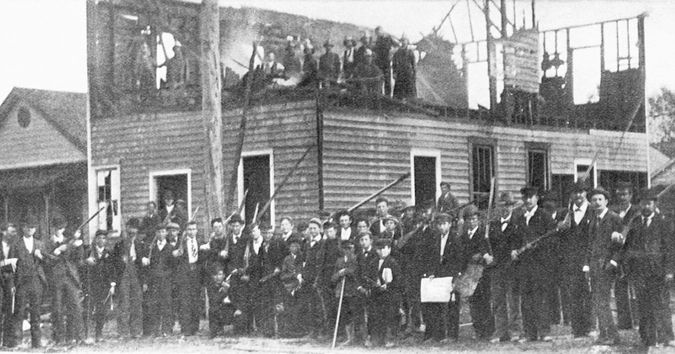 A mob of white supremacists pose with their rifles in front of the demolished printing press of the Daily Record, an African American newspaper during the Wilmington Insurrection. Some members stand atop the burnt out second story of the building.