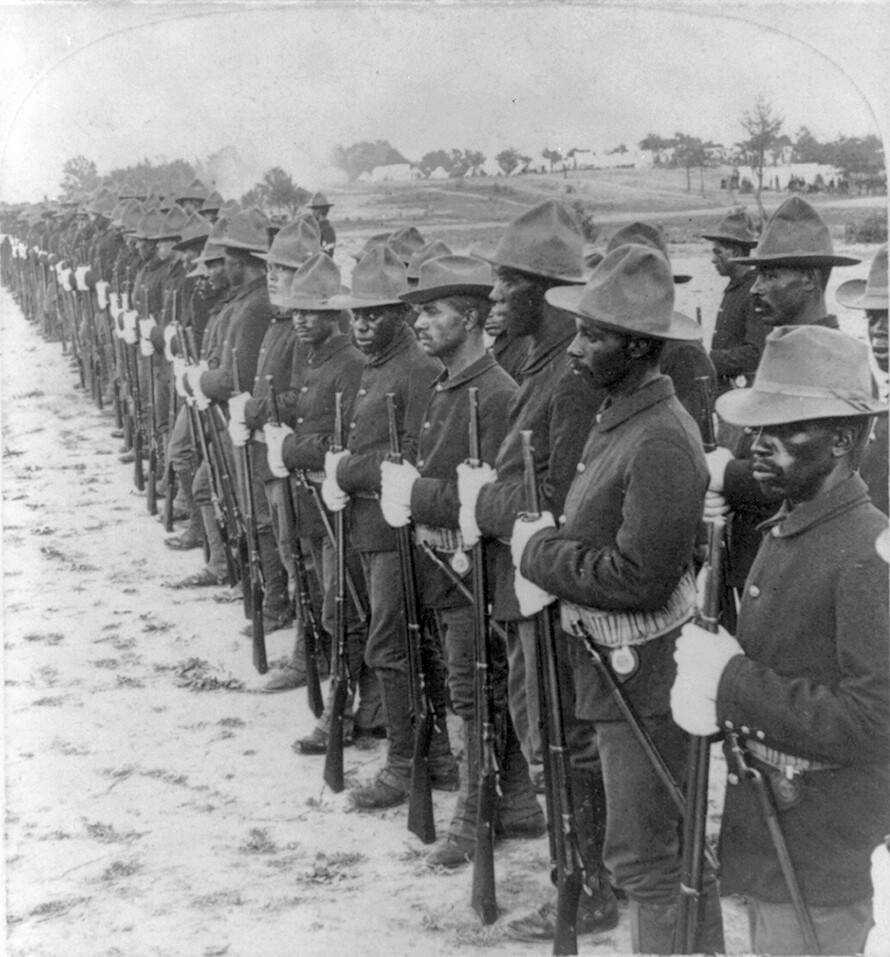 African American troops in Cuba stand in formation wearing old wool uniforms unsuited to Cuba"s tropical heat. Each man is holding a rifle in front of himself.