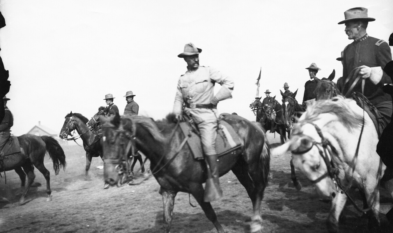With one hand on his hip, Colonel Roosevelt rides with the Rough Riders in Cuba. Several men on Horseback surround him.