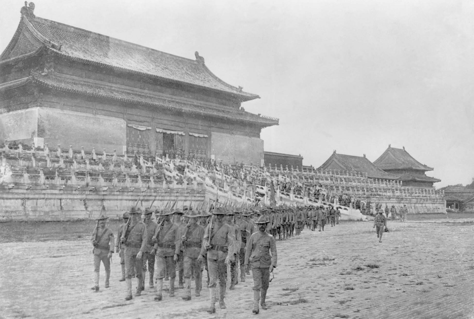 A black and white photo shows a long line of U.S. troops in rank marching in the Forbidden Palace, the imperial palace in the Chinese capital of Peking, after quelling the Boxer Rebellion.