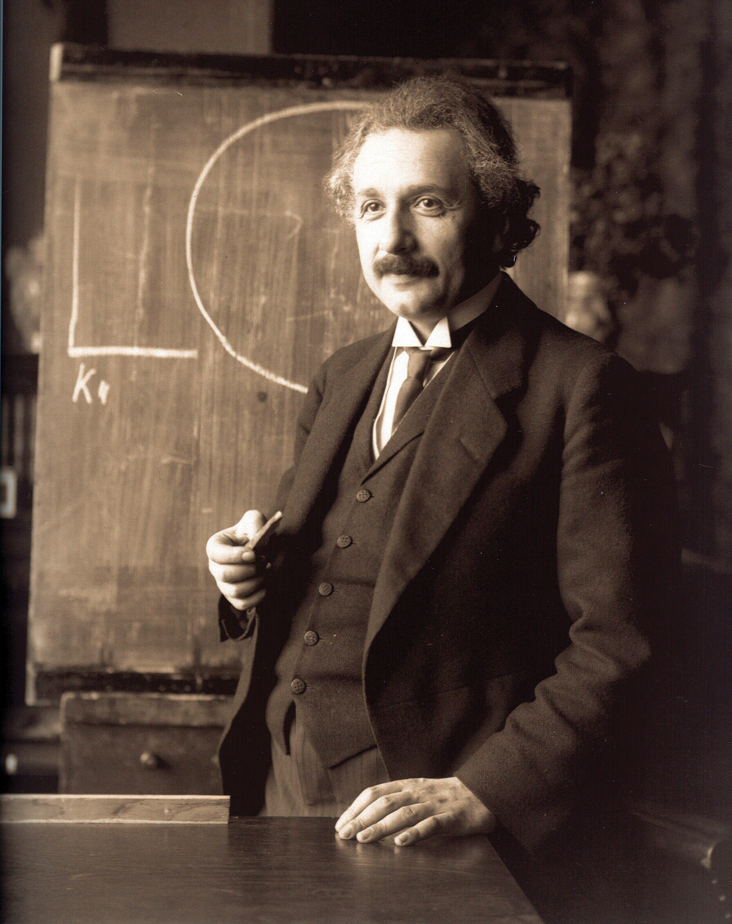 A sepia photograph of a man in a suit, posing in front of a chalkboard.