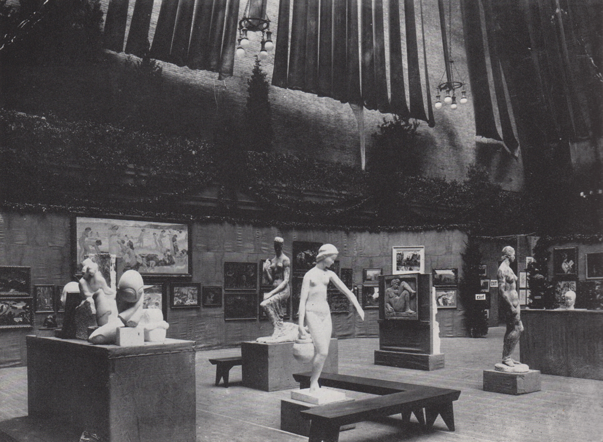 A black-and-white photograph of a large exhibition room containing sculptures, carvings, and paintings.