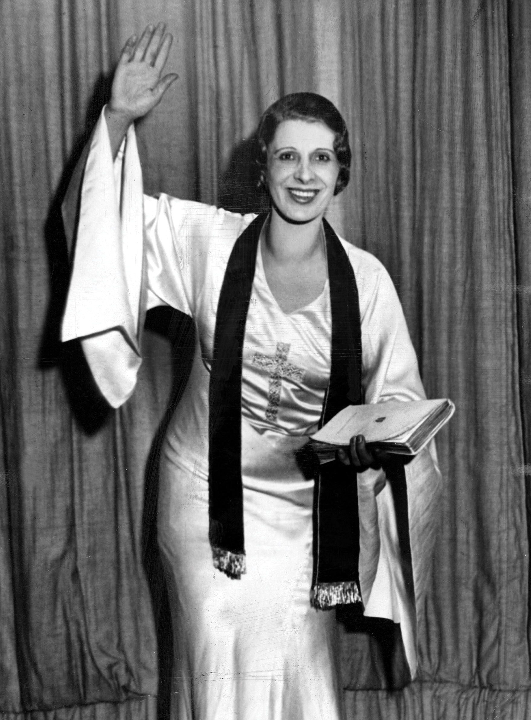 A black-and-white photograph of a smiling woman, waving with one hand and holding a book in the other.