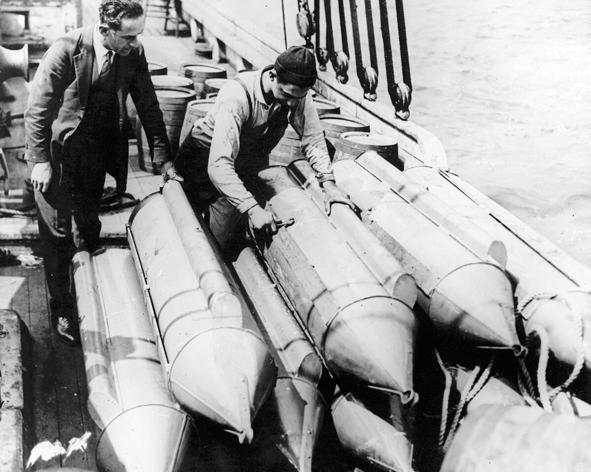 A black-and-white photograph of two men standing on a boat near a stacked pile of torpedoes.