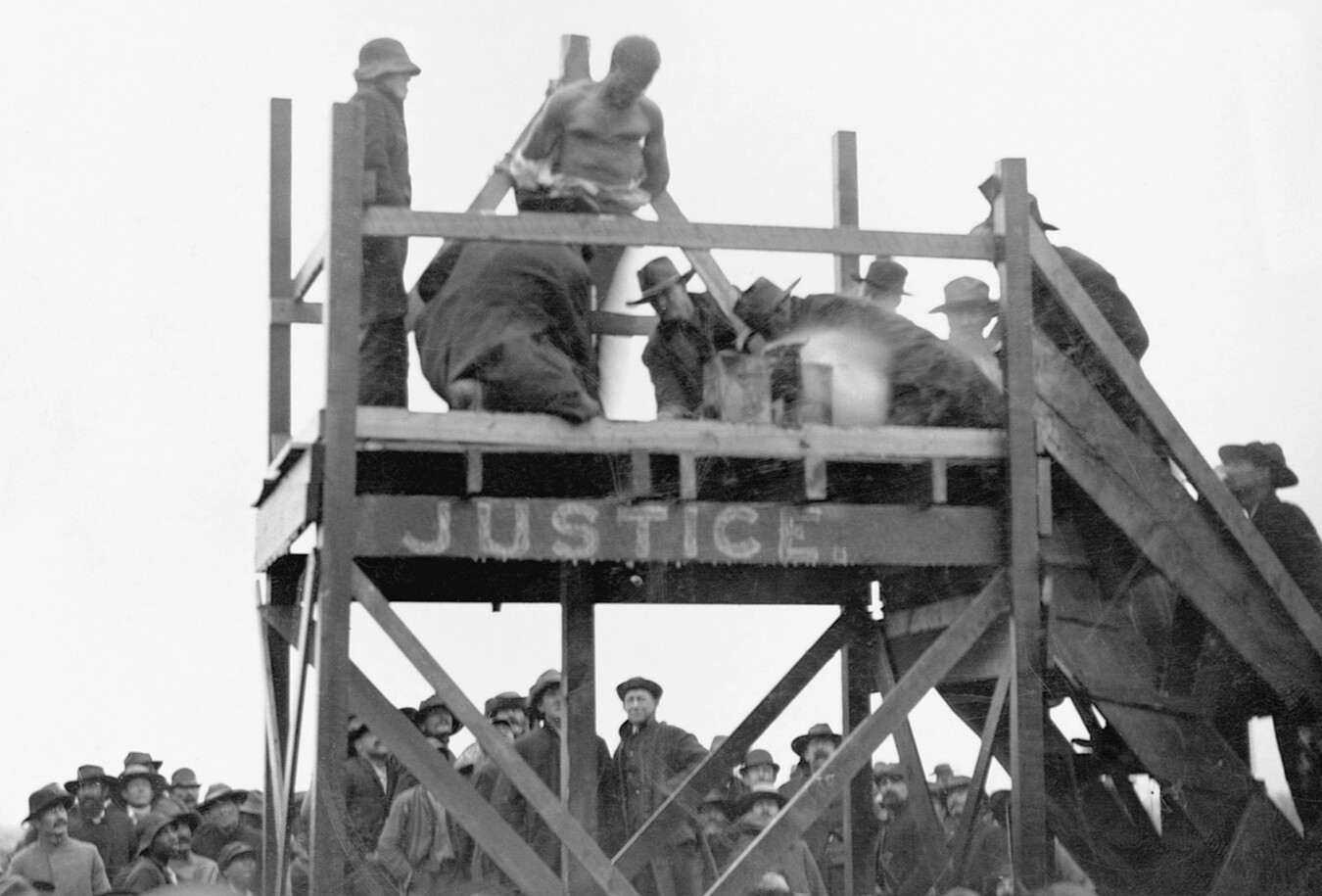 A crowd of spectators watch a white men surrounding a Black prisoner on a platform.