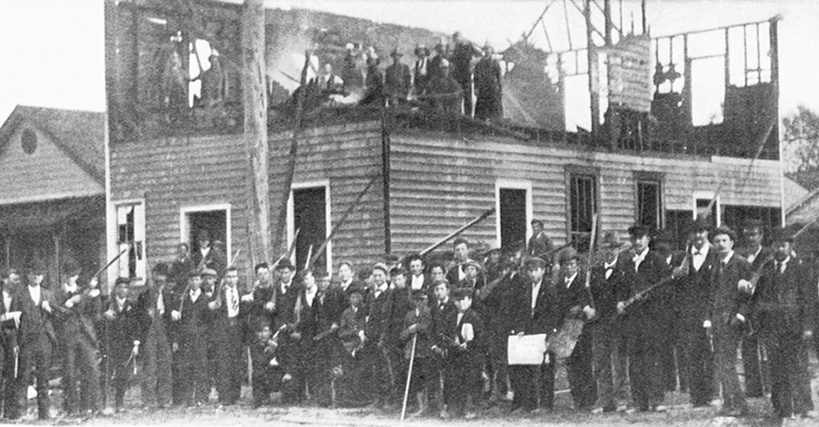 A large crowd of White men with rifles stand in front and on top of a wrecked building.