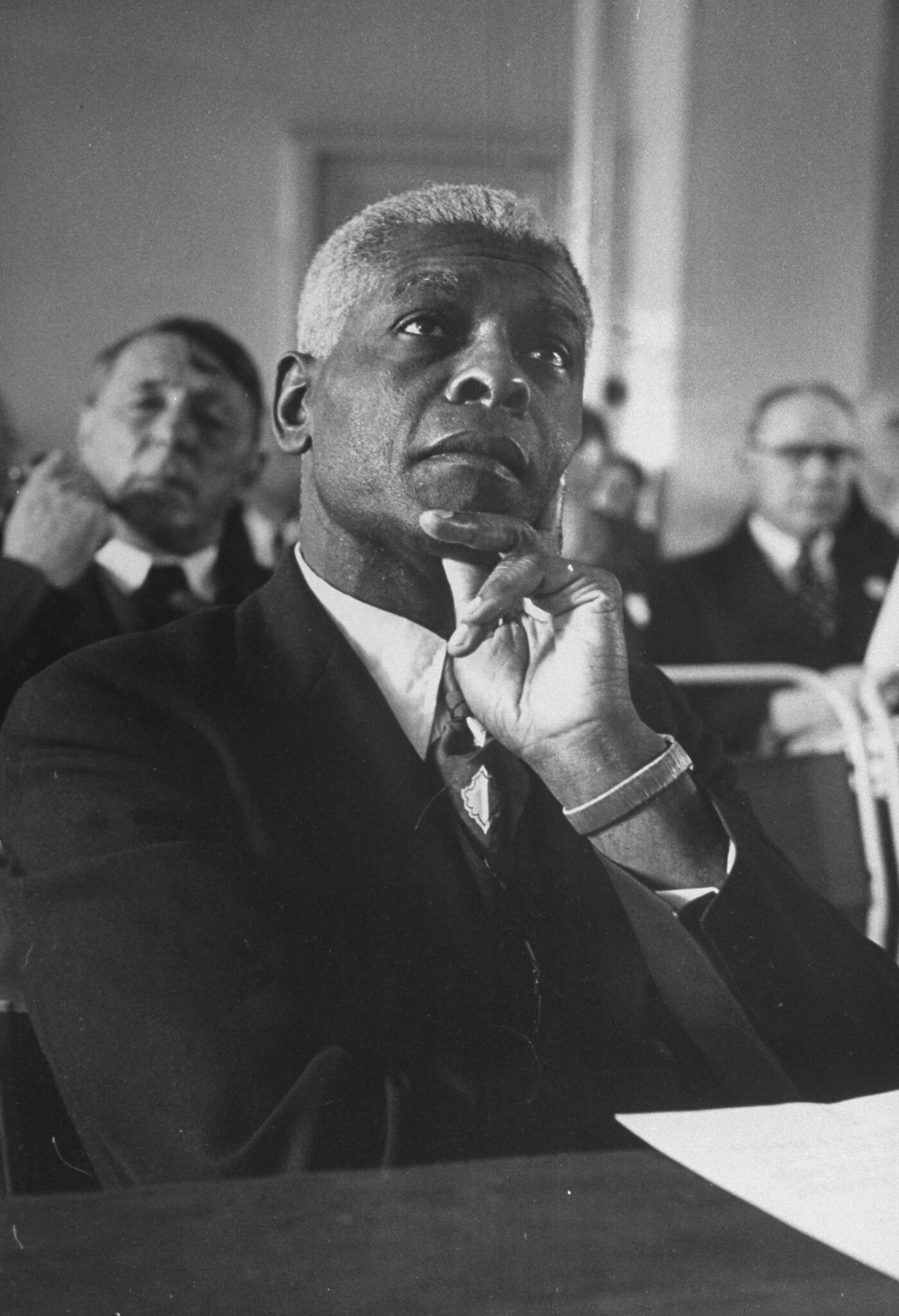 A photo of a man in a suit and tie sitting in a room and resting his chin on his hand.