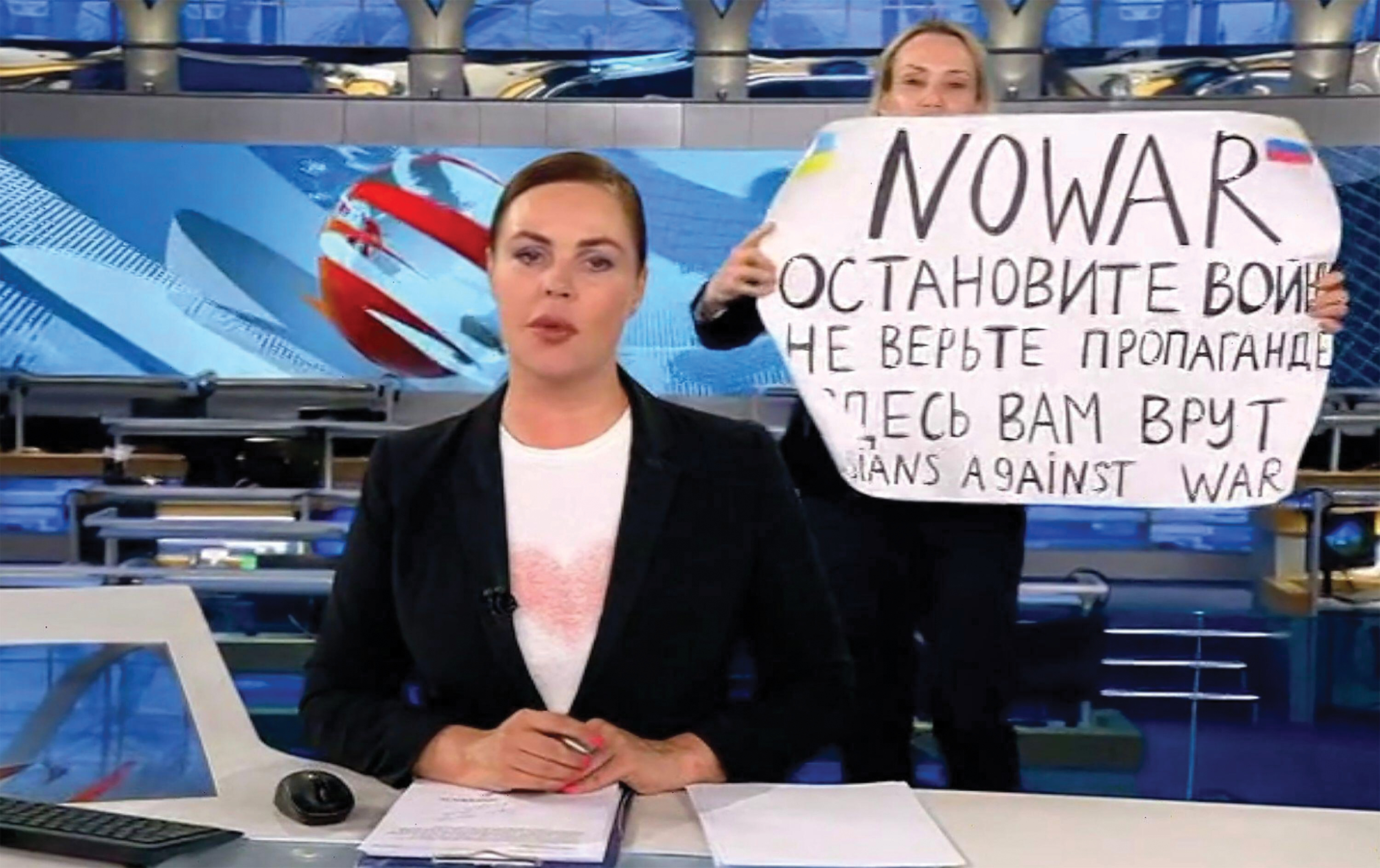 A photo of a news reporter sitting at a desk in front of news screens.