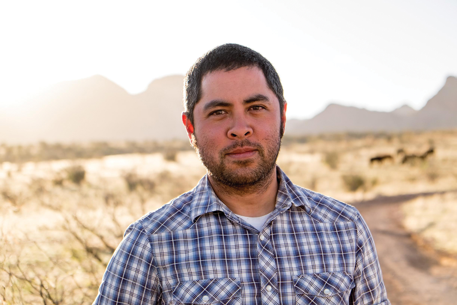 A photo of Jason De León, a man with short hair, short facial hair, and a plaid shirt, standing on a path in a desert.