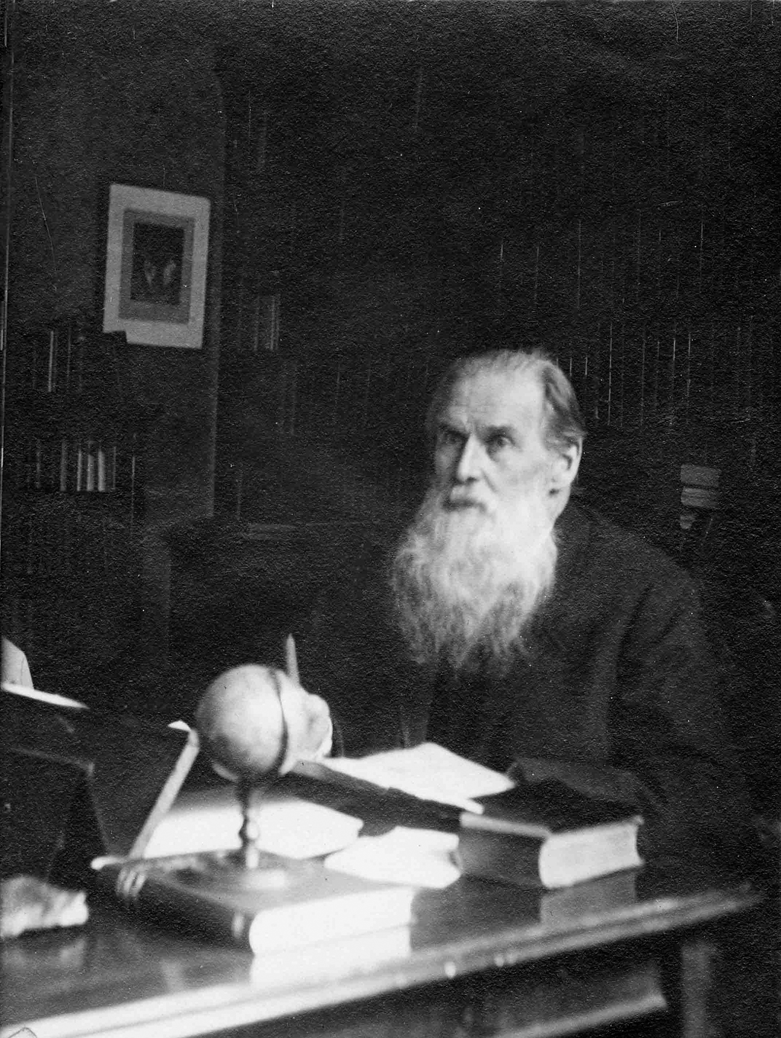 A grayscale photo Edward Burnett Tylor, a white man with a long, white beard sitting at a desk covered with books.