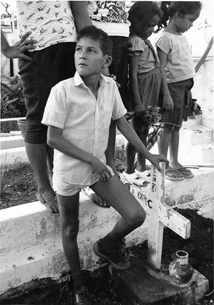 A black and white image of a young boy standing with his hands placed on a wooden cross marking a grave.