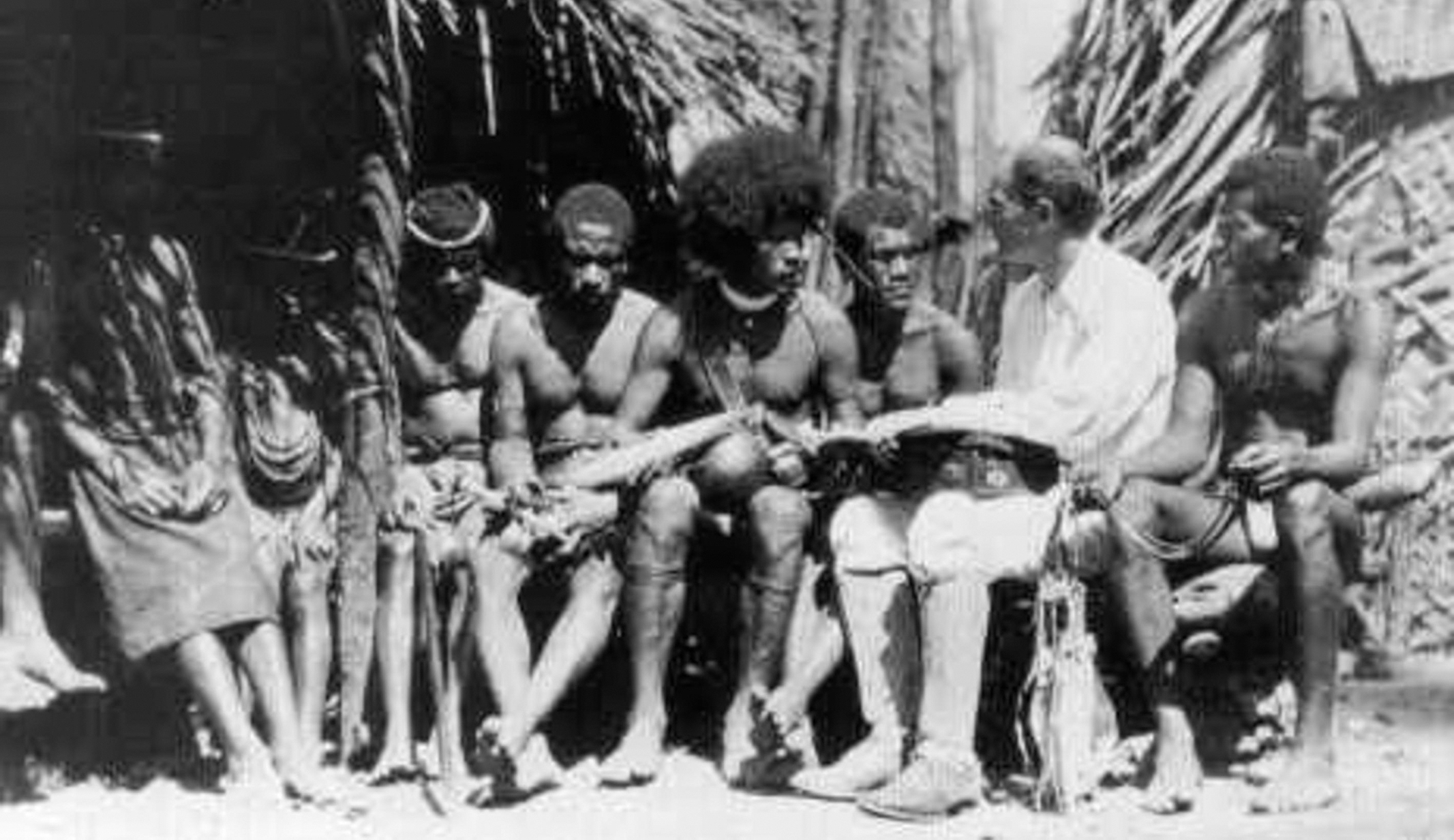 A black and white photo of a white man wearing knickers and high boots talking with a group of people in loincloths.