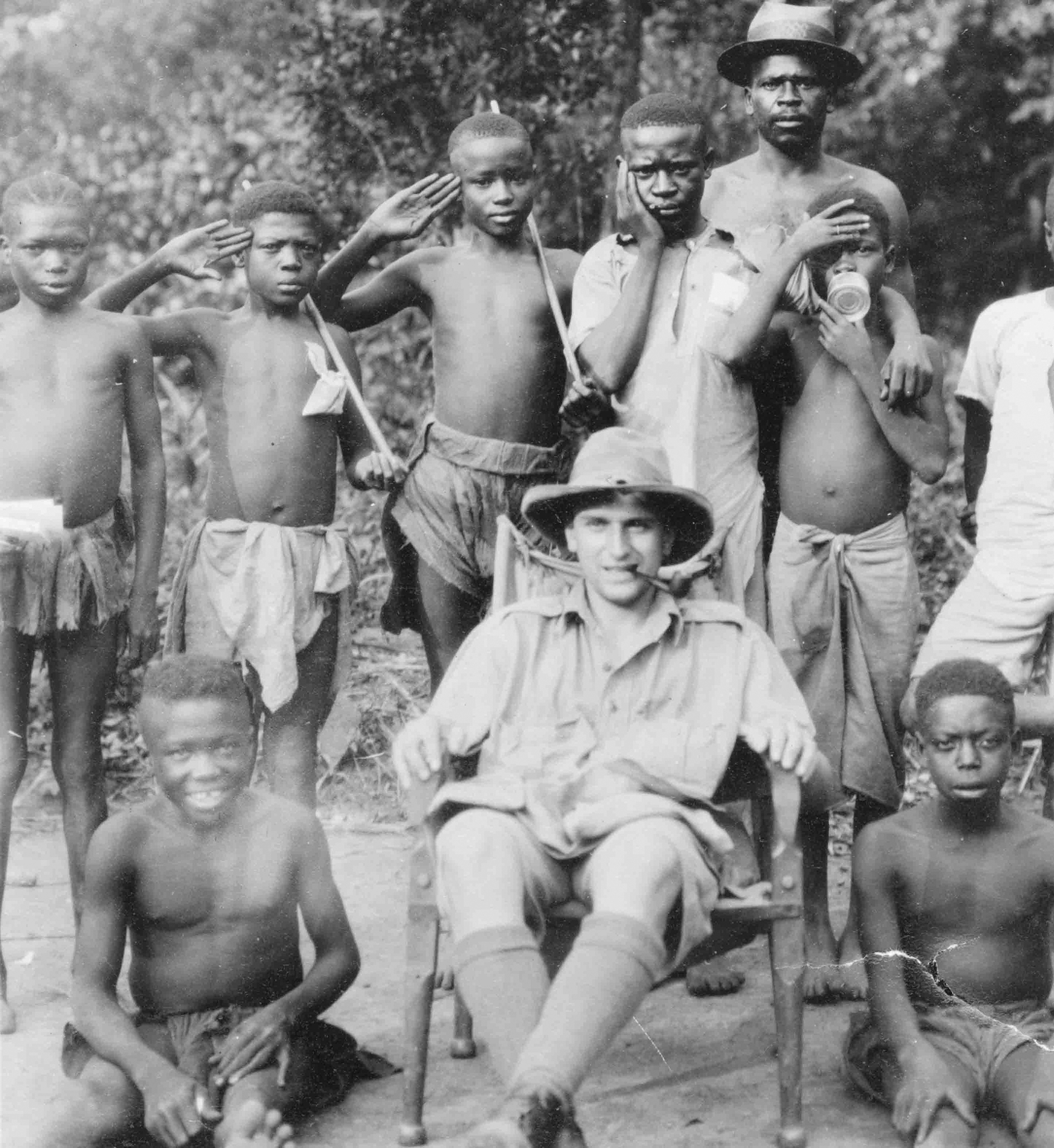 A black and white photo of a white man in a wide-brimmed hat sitting in a chair surrounded by Nuer people in loincloths.