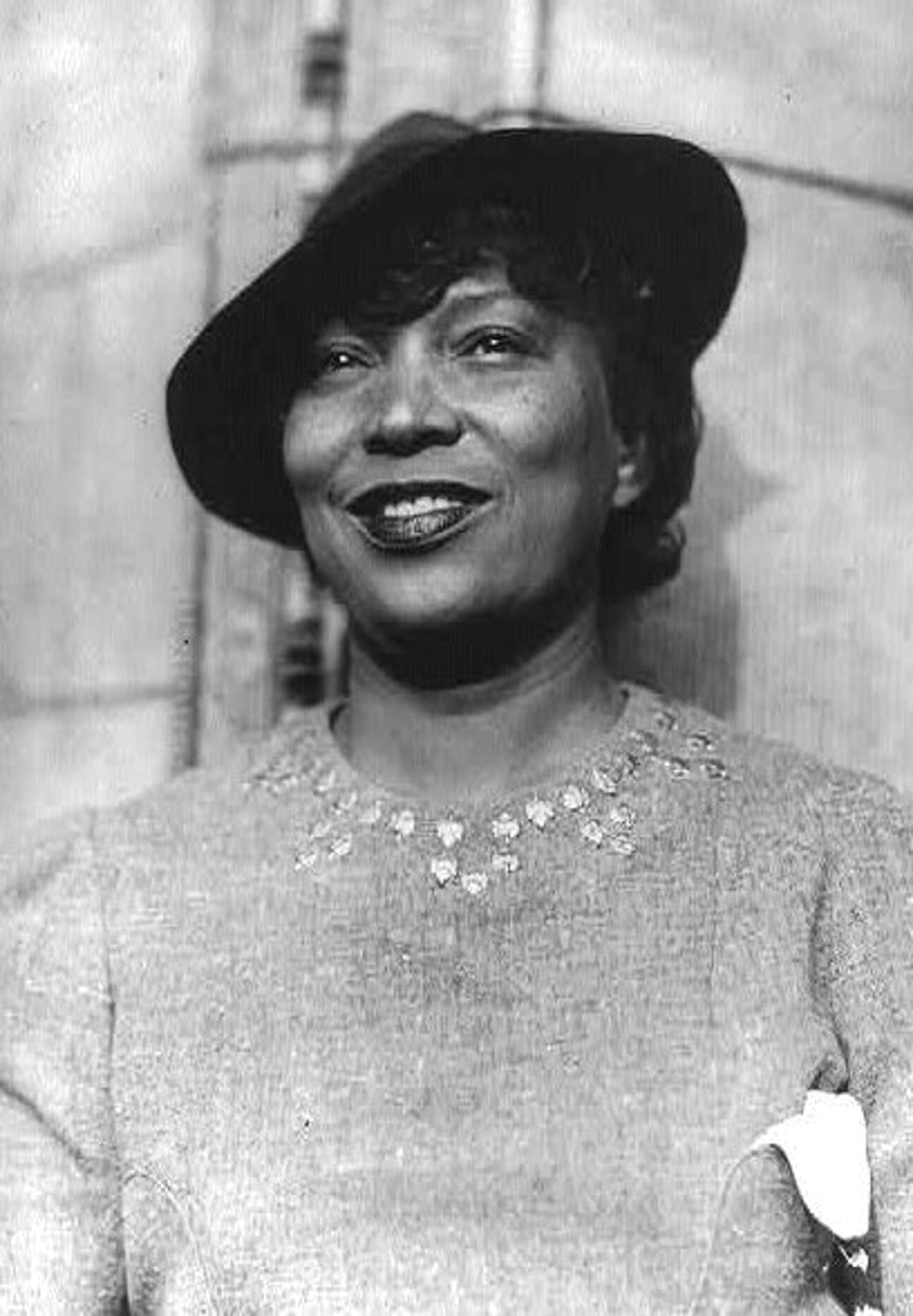 A black and white photo of Zora Neale Hurston, a Black woman, wearing a hat and smilling.