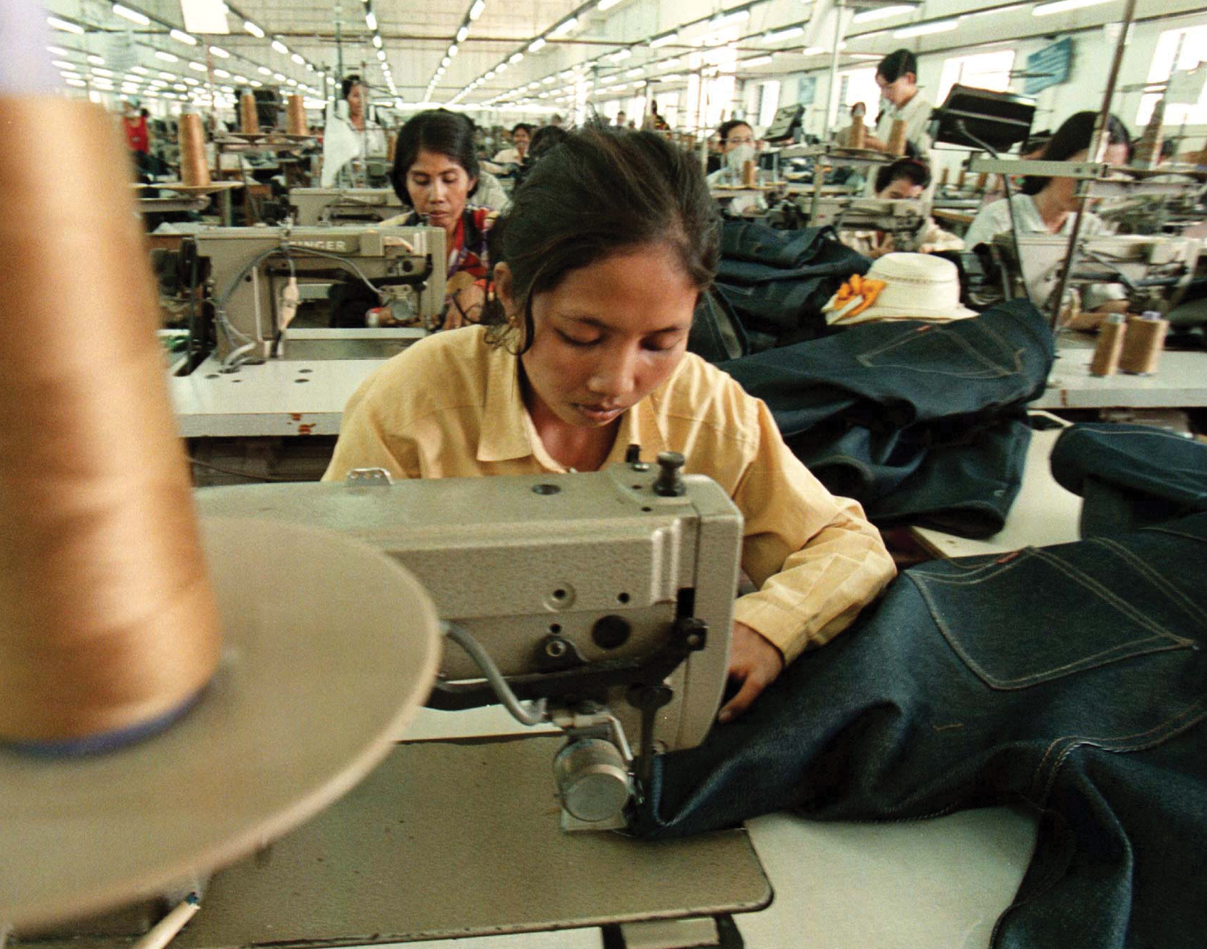 A woman sits on a production floor sewing blue jeans with other women also working.
