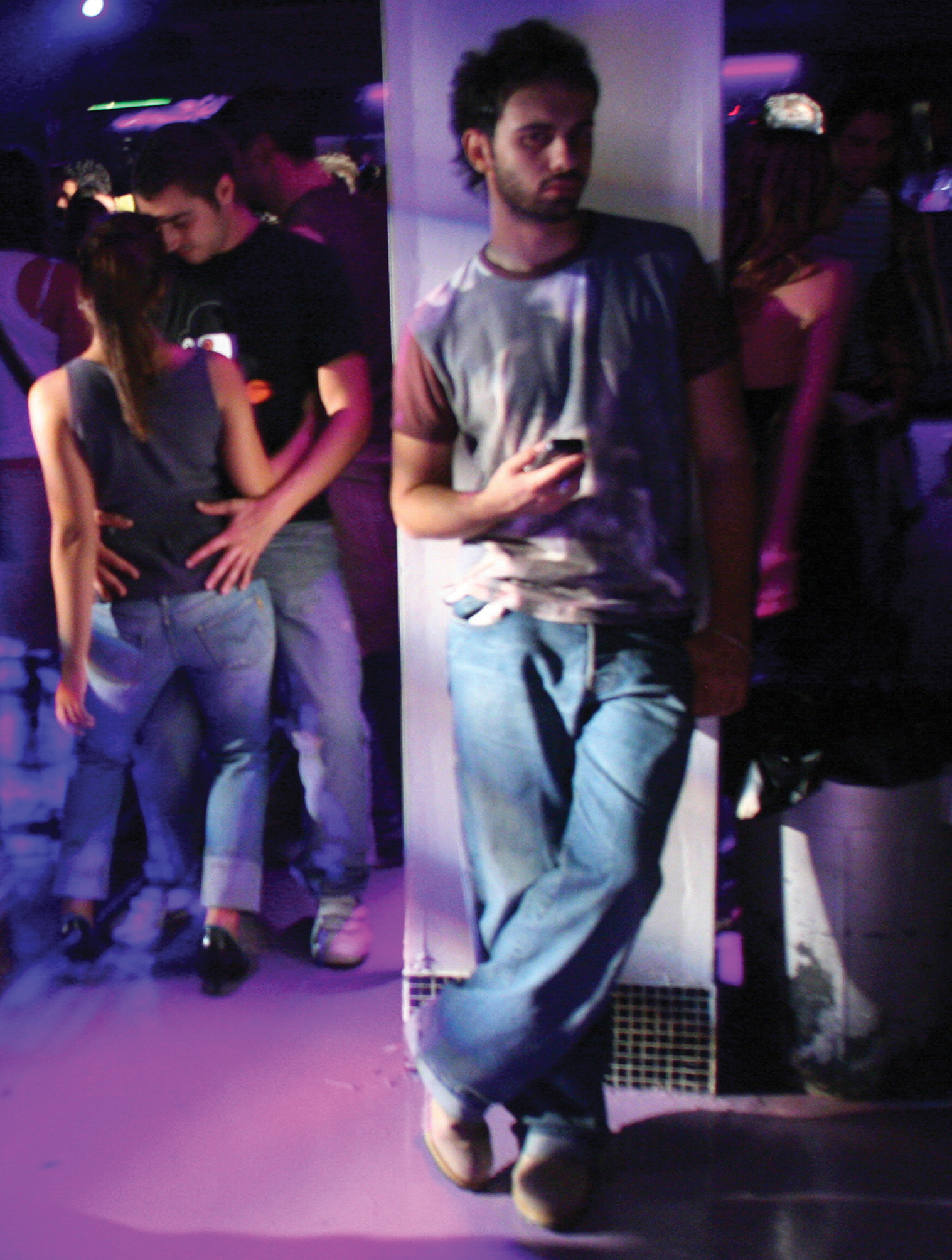 A man in a nightclub wearing a T-shirt and baggy jeans looks at a cellphone while leaning against a pillar.