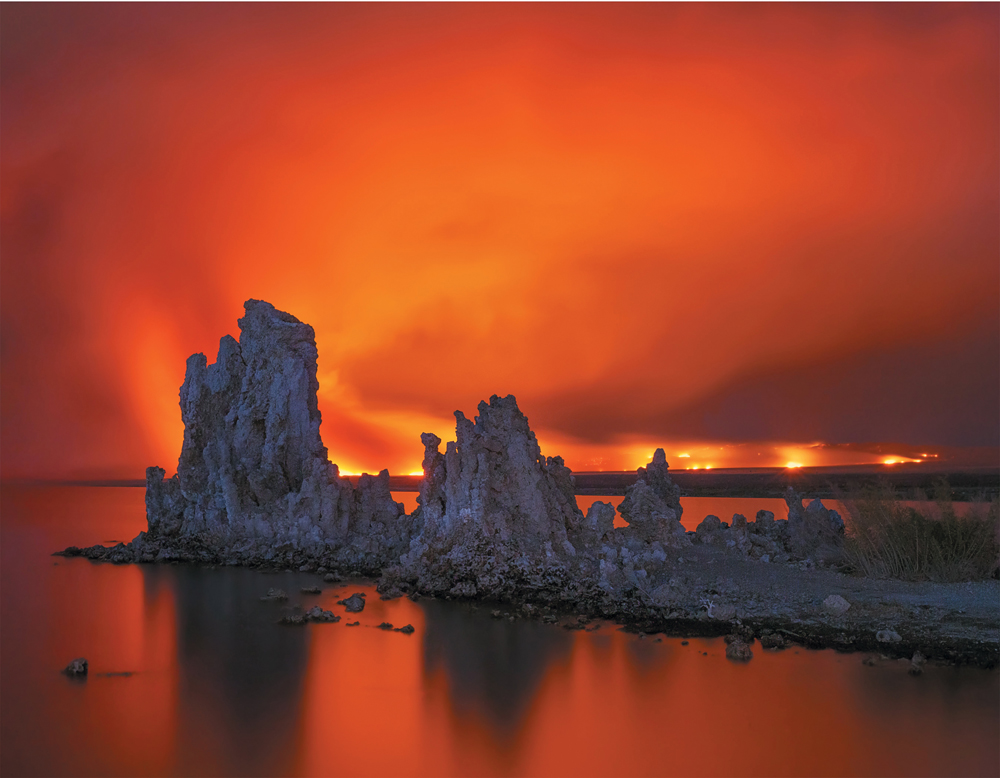 A photo of Mono Lake at sunset. Behind a jagged outcropping of rock projecting up from the water is a smoky coastline and a bright red sky reflecting off the lake’s surface.
