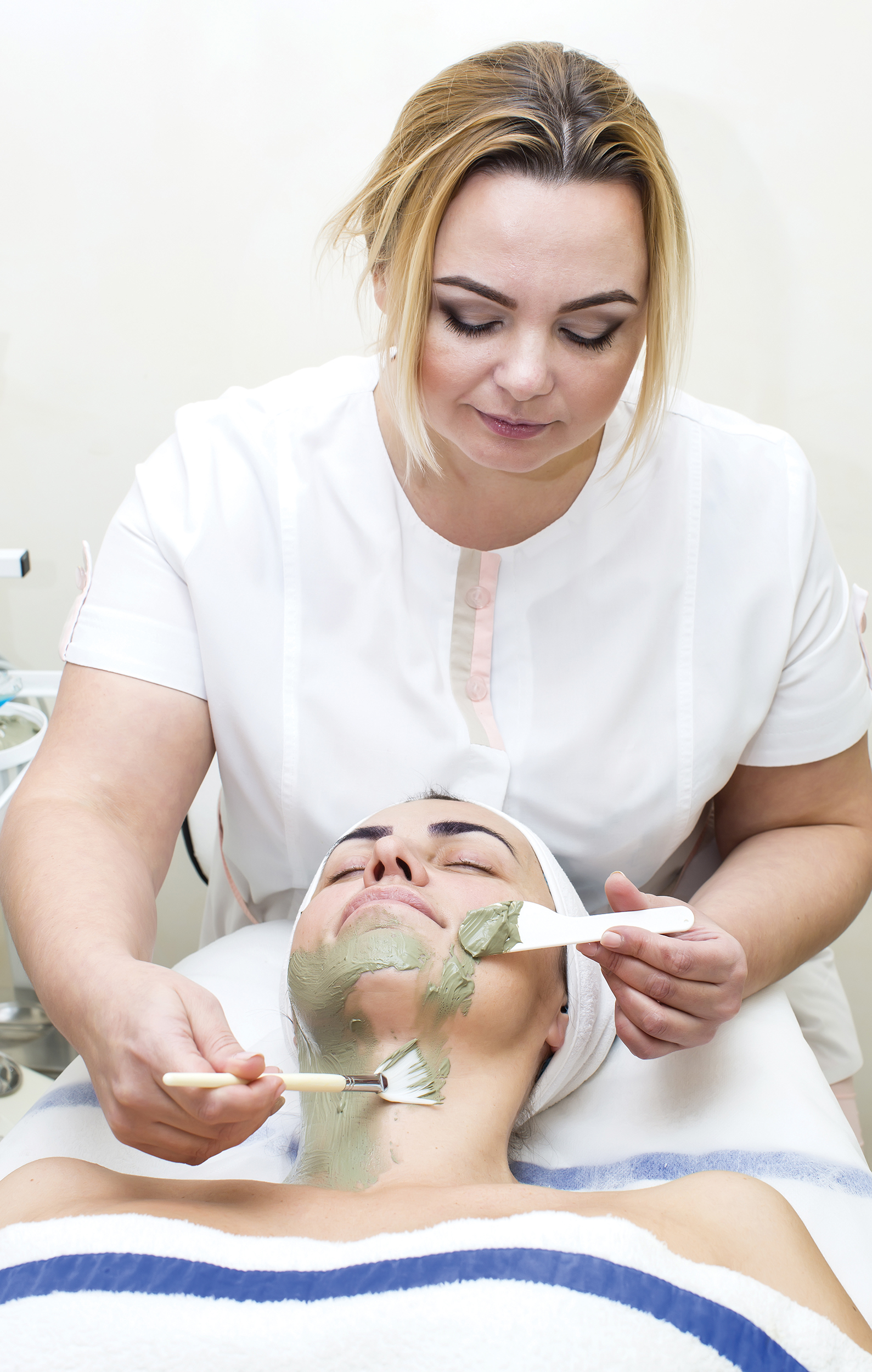 A woman lying down receives a spa facial treatment from another woman dressed in white.