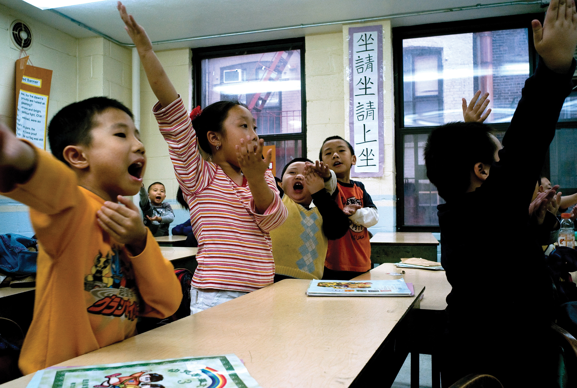 A group of children stand singing and making hand gestures in a classroom.