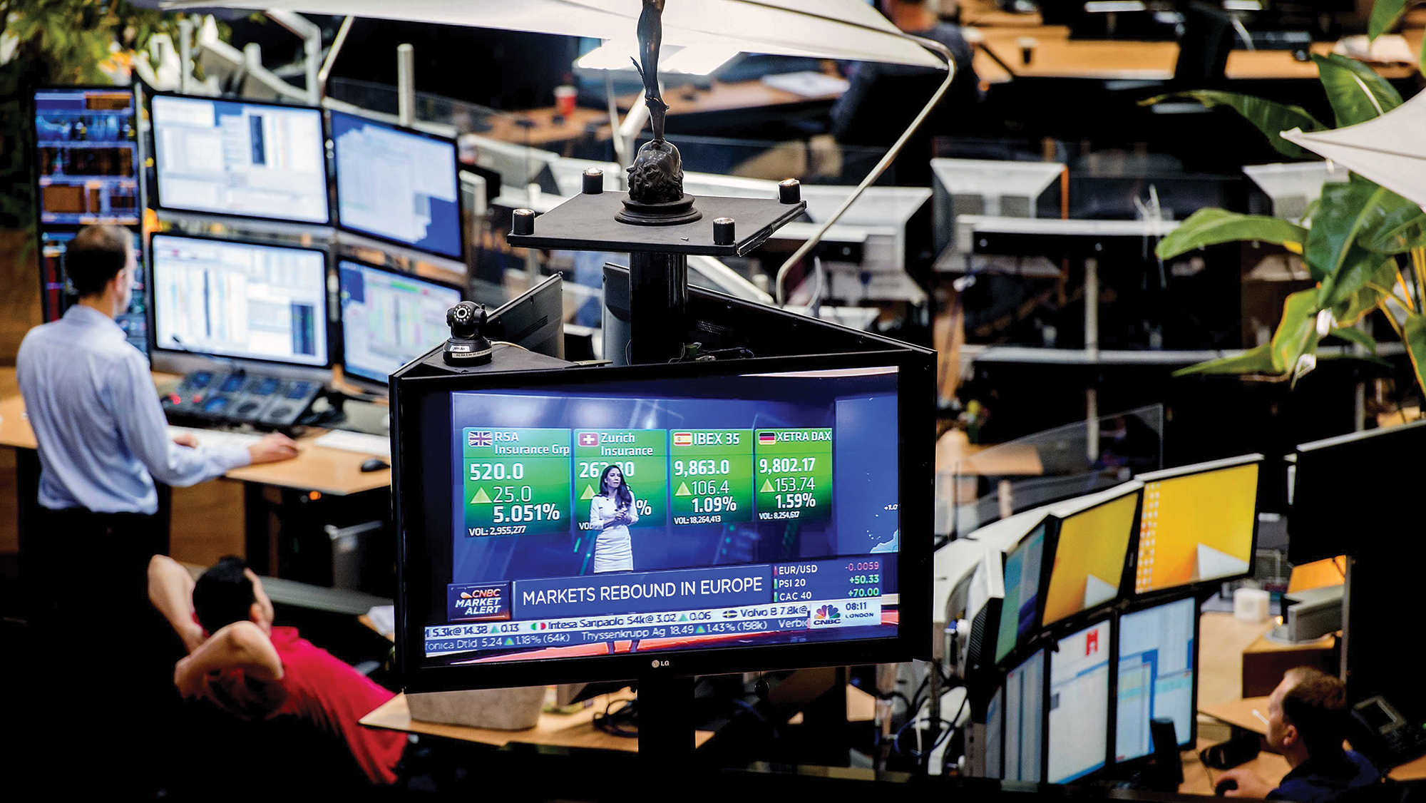 Men sit and stand looking at numerous computer screens that show stock prices.