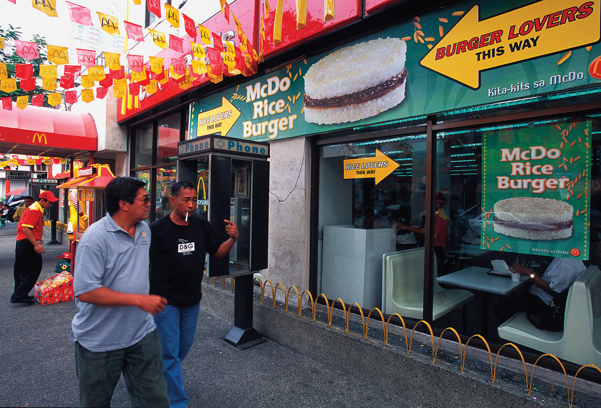 Two people walking past a McDonald’s restaurant with numerous signs advertising the McDo Rice burger.