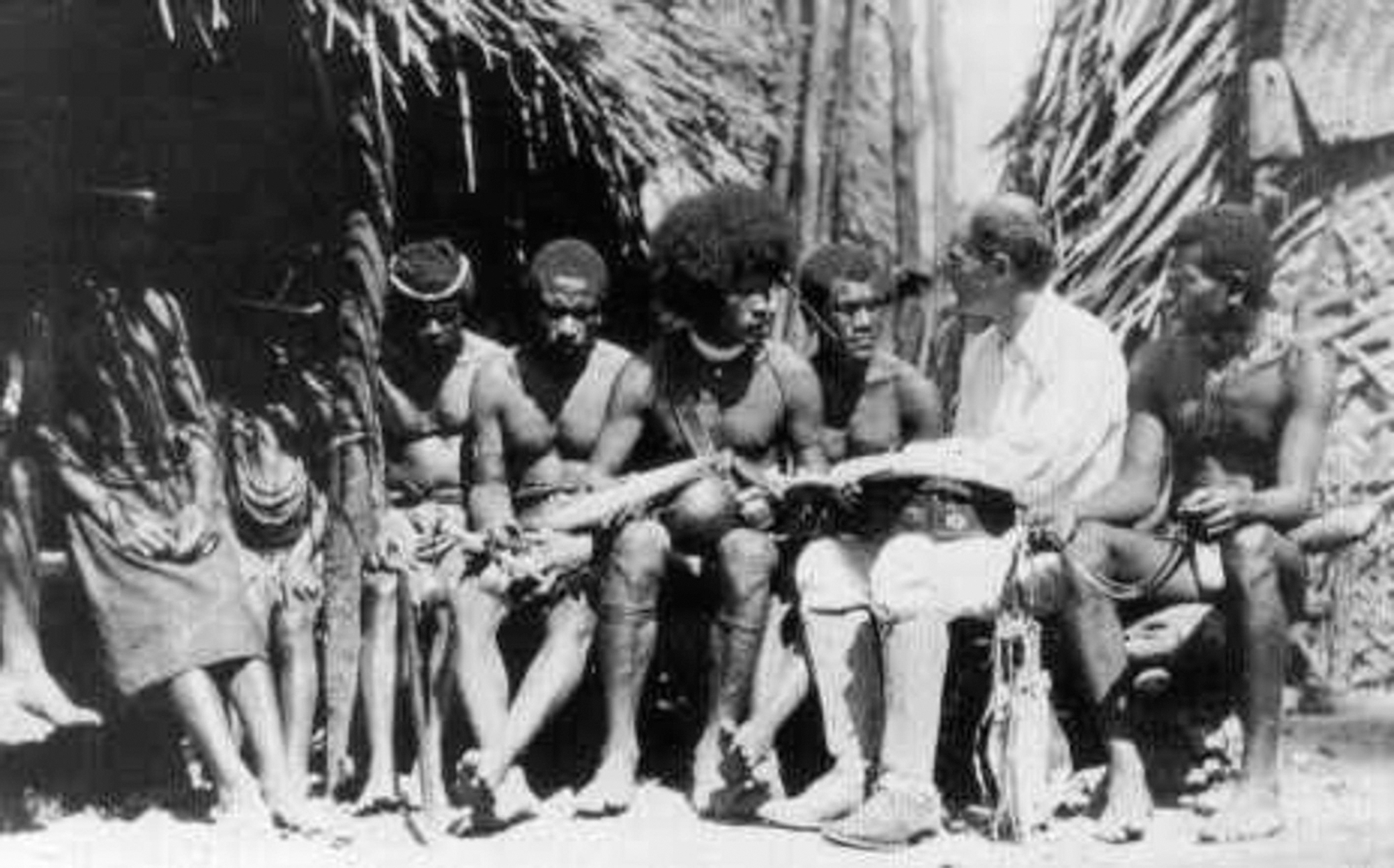 A black and white photo of a white man wearing knickers and high boots talking with a group of people in loincloths.