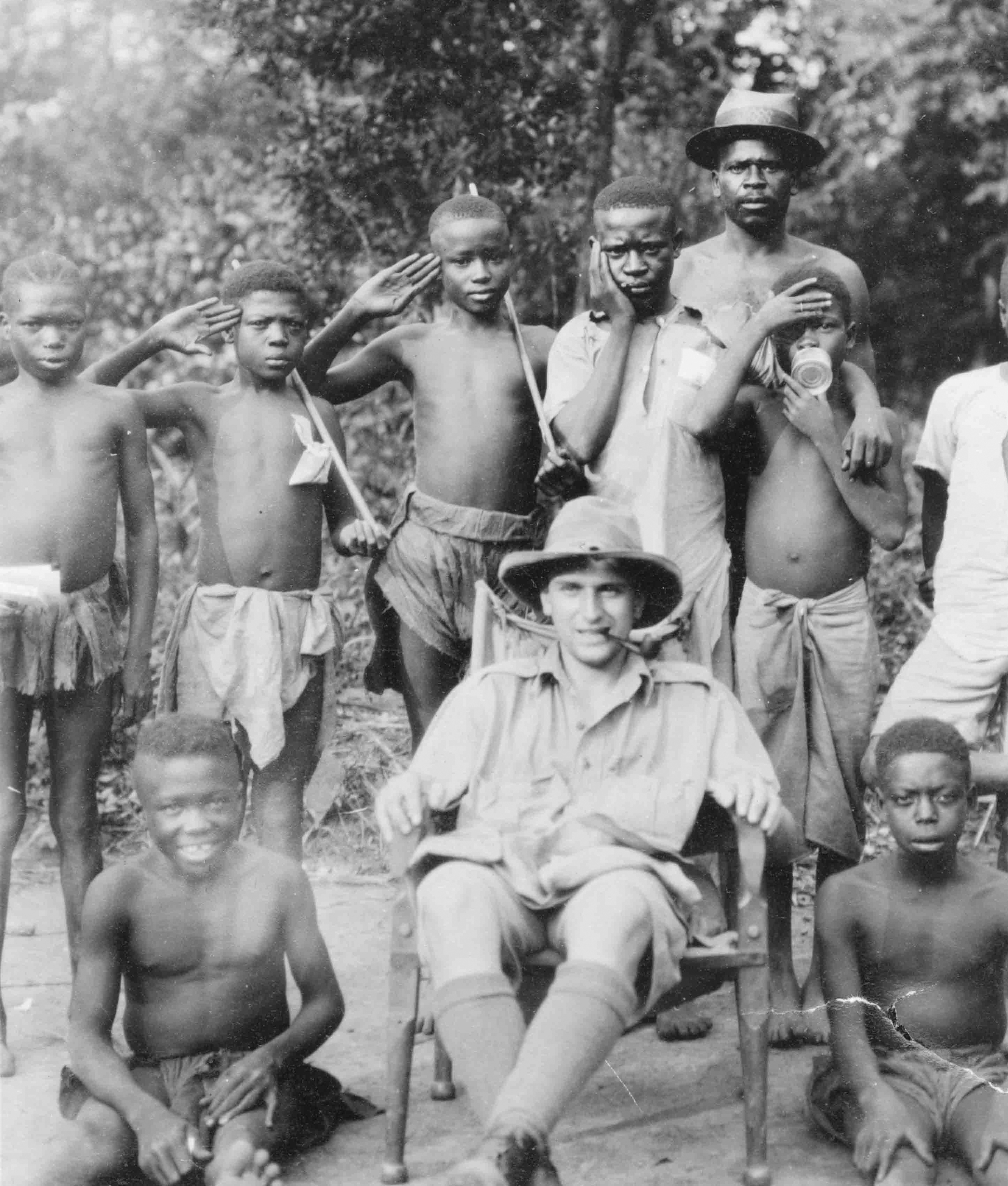 A black and white photo of a white man in a wide-brimmed hat sitting in a chair surrounded by Nuer people in loincloths.