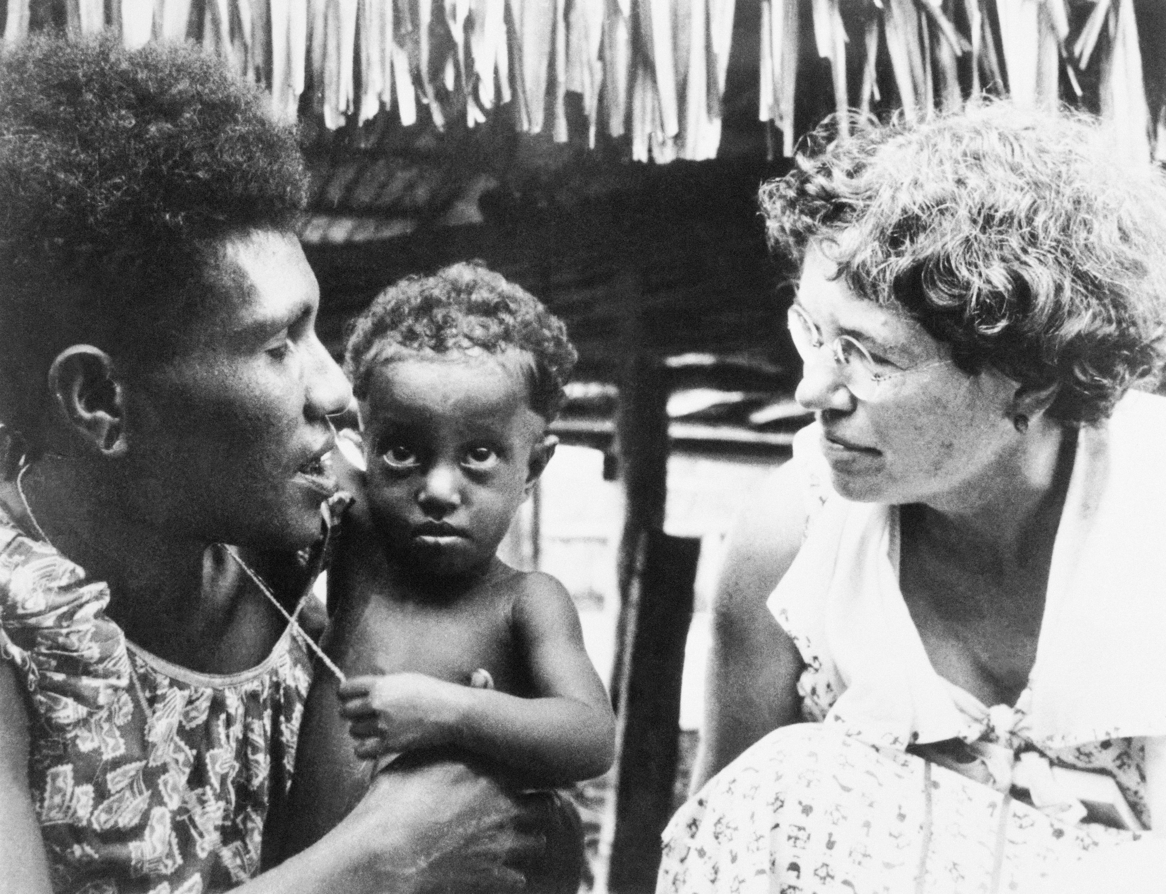 A black and white photo of Margaret Mead, a white woman, wearing glasses and speaking to a woman holding a baby.
