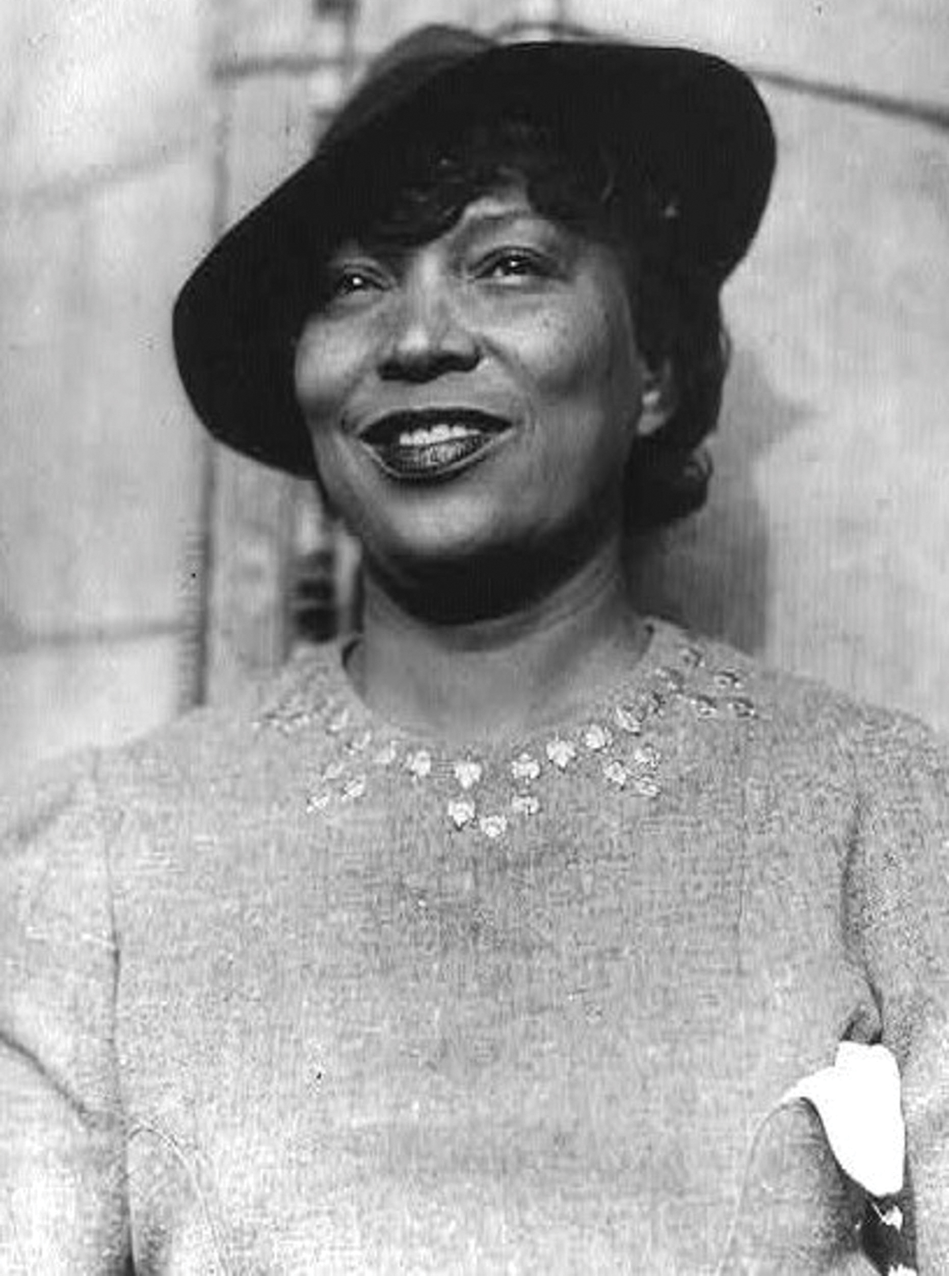 A black and white photo of Zora Neale Hurston, a Black woman, wearing a hat and smilling.