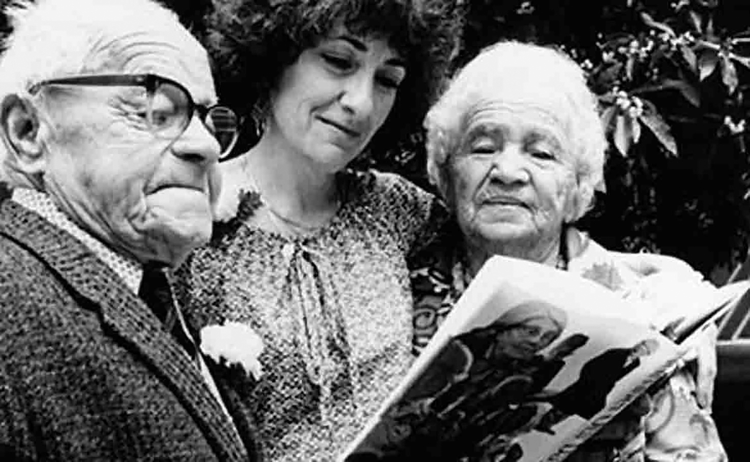 A black and white photo of a young white woman standing between two senior citizens looking at a book.
