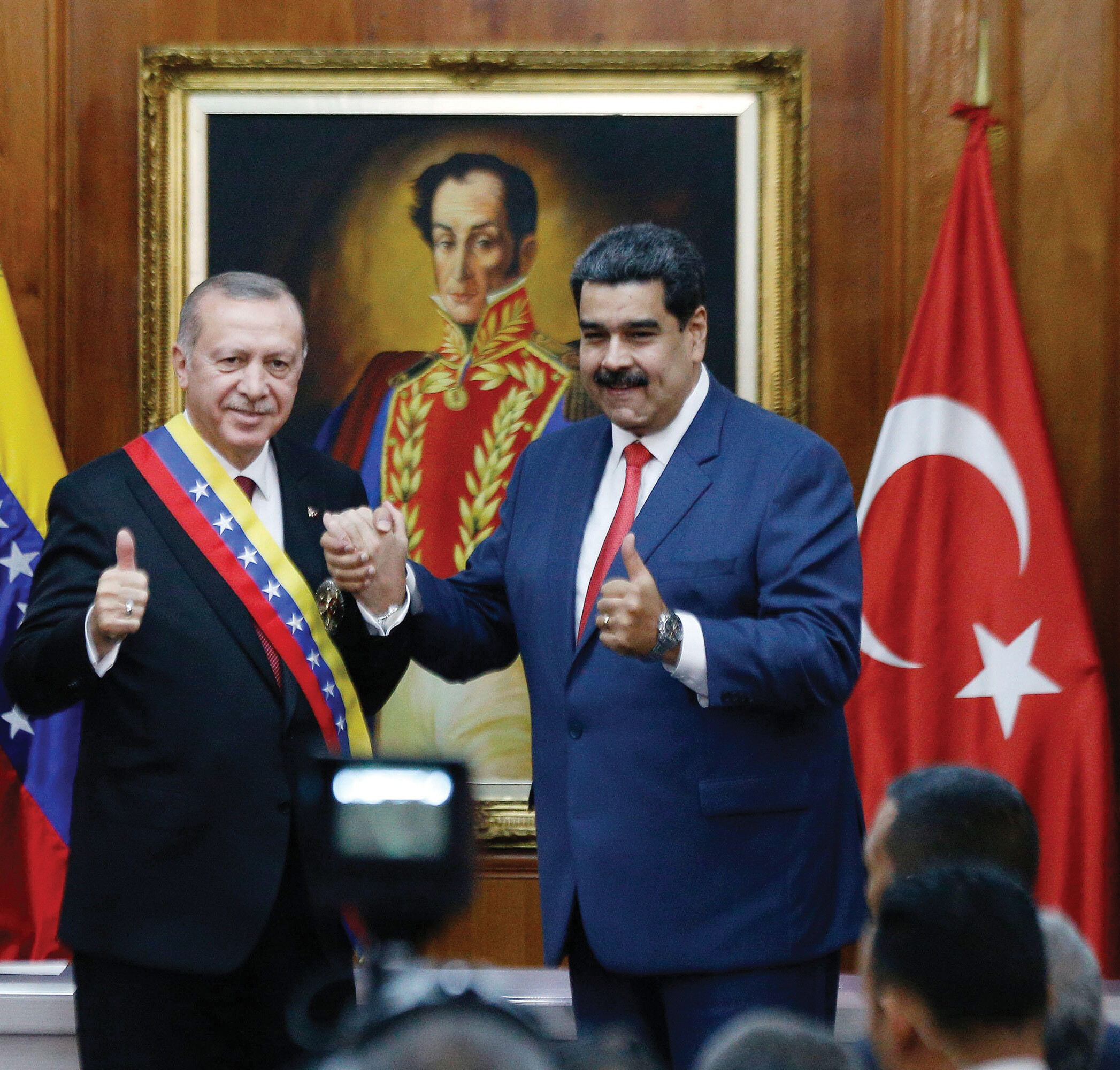 Two men stand together in front of Venezuelan and Turkish flags. They are grasping hands while giving the thumbs up sign with their other hand.