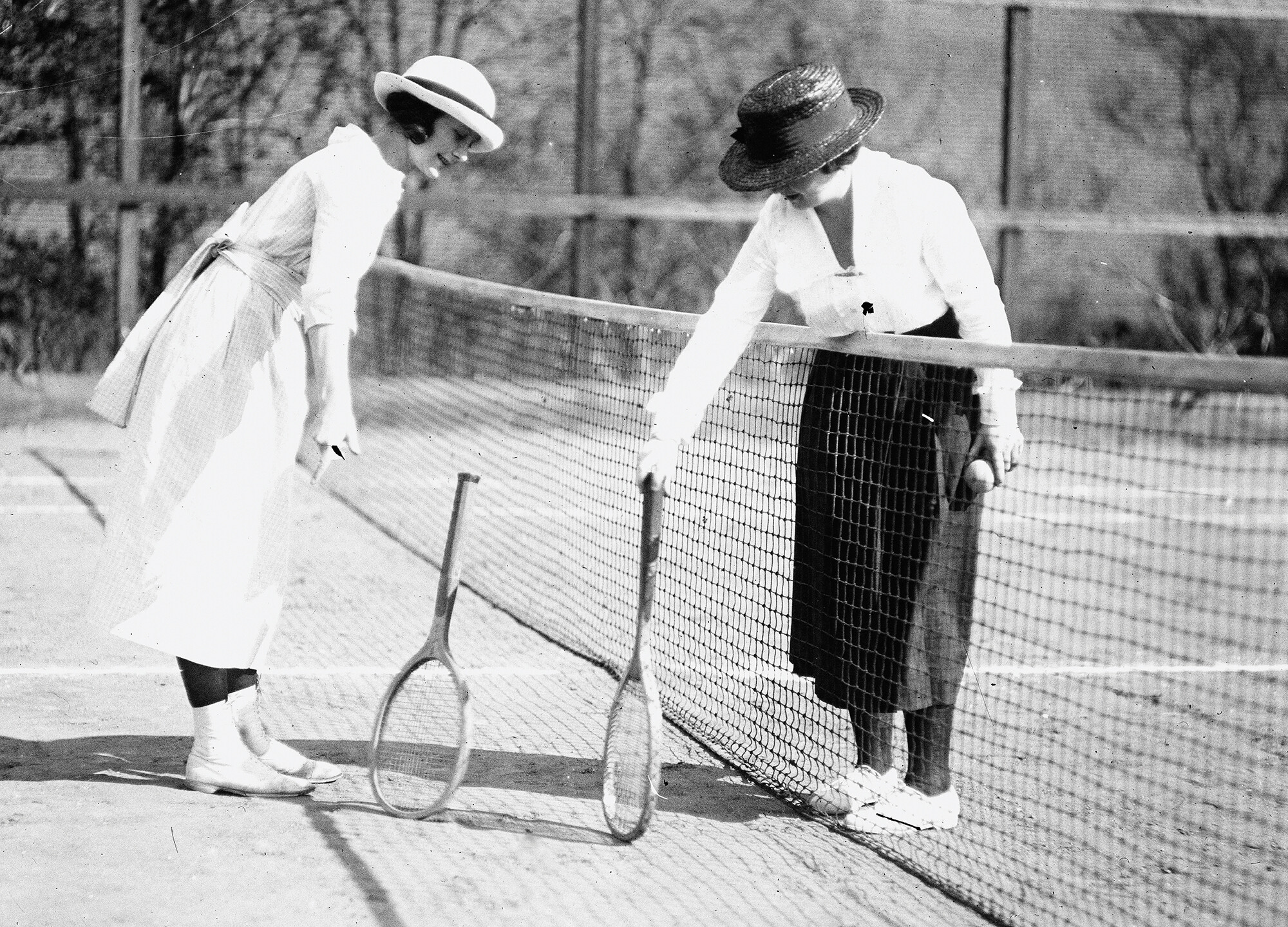A black and white photo of two women on either side of a tennis net attempting to balance their rackets on the rackets’ round ends. One They both wear long-sleeved blouses, long skirts, and hats.