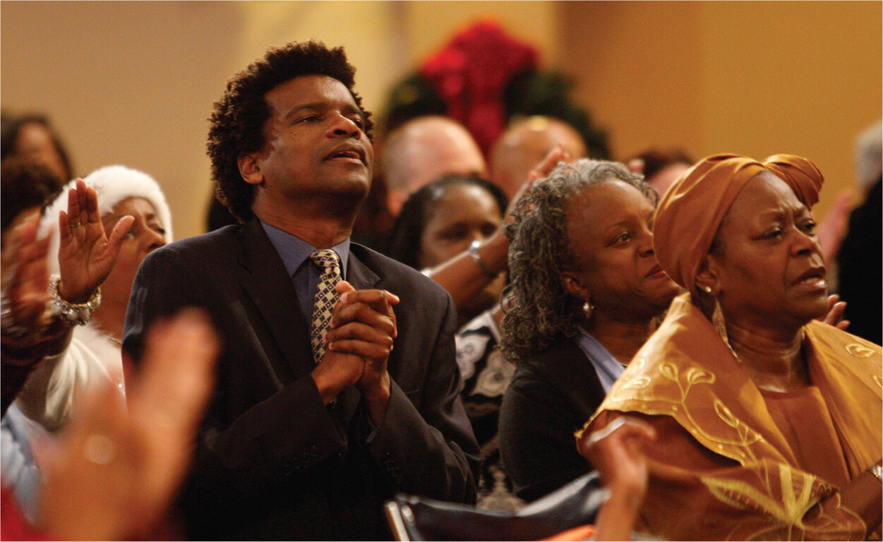 A group of Black congregants seated in rows look ahead. Some clap their hands, while others raise them up and forward, palms open. One man clasps his hands together.