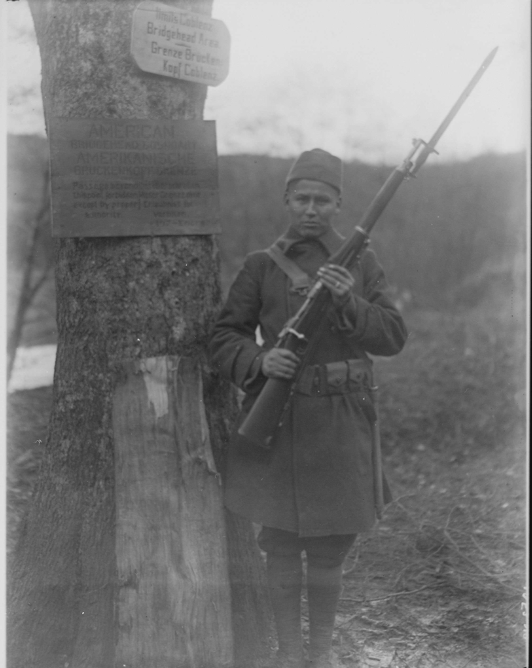 A black-and-white photo of George Miner standing guard with a bayonet next to a tree that has multiple signs.