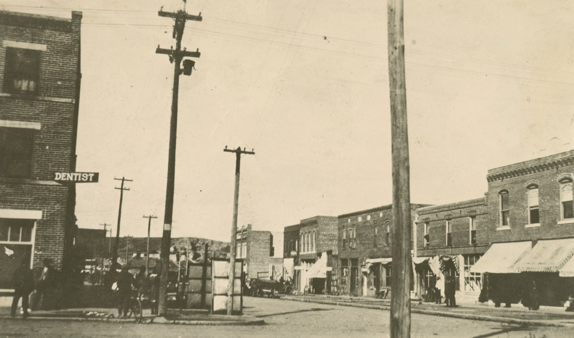 A sepia photo of an intersection with electric poles and stone buildings. A dentist sign hangs from one of the buildings.