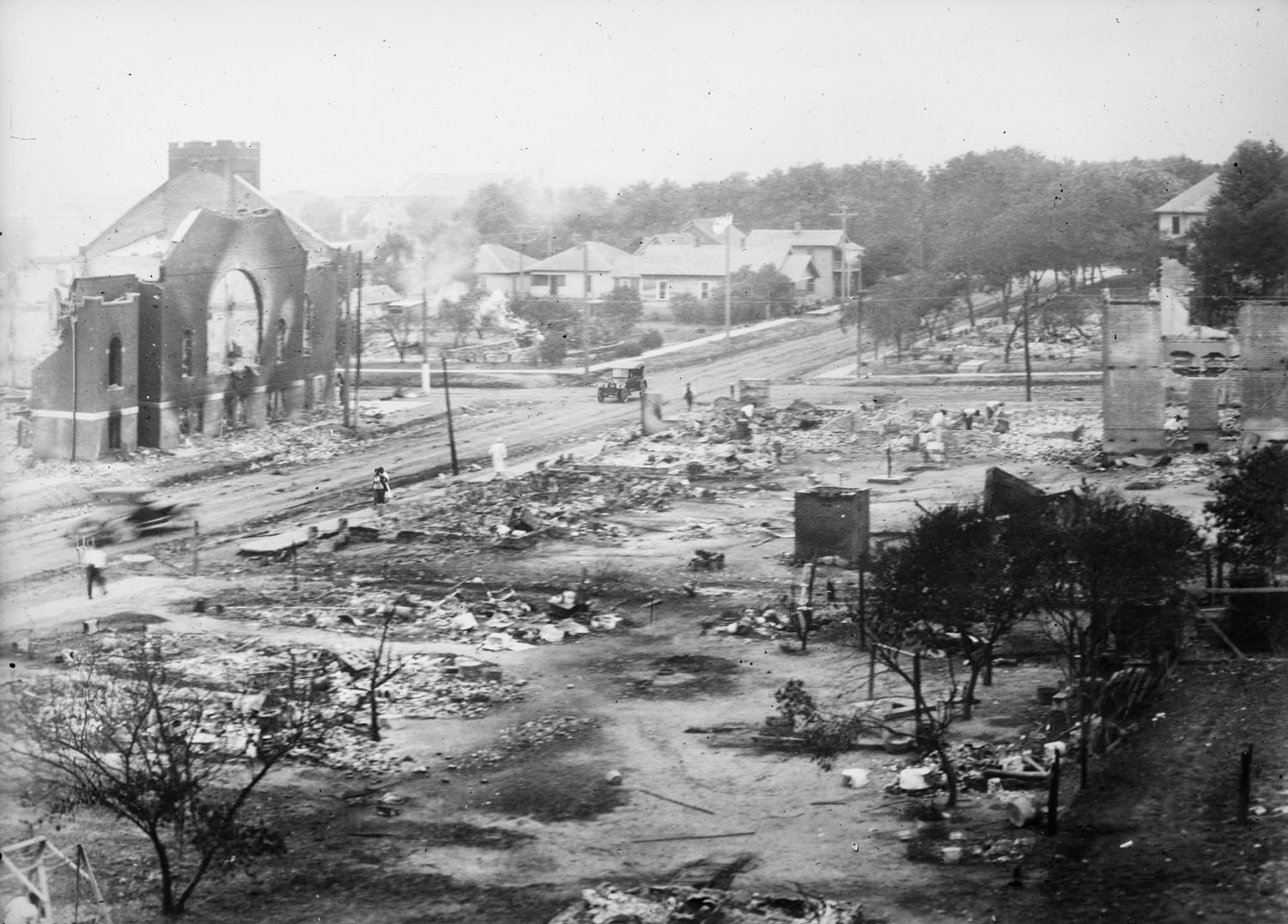 A black-and-white photo of an aerial view of rubble of buildings. People walk through the rubble.