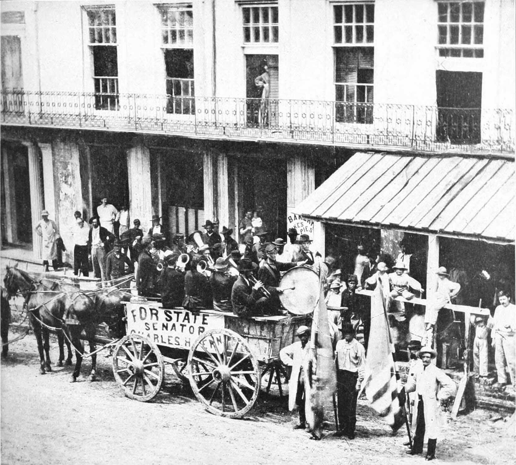 A black-and-white photo of people in a horse cart and playing band instruments. A banner on the cart reads, For state senator. A man standing next to the cart holds the American Flag.