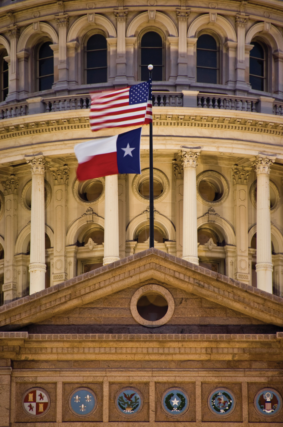 Two flags flying in front of the Texas state capitol: the American flag and the Texas state flag.