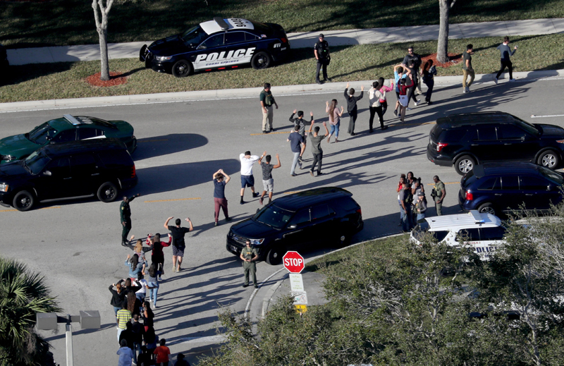 A photo shows a queue of teenagers walking with their hands raised. A Police car is parked on the roadside.