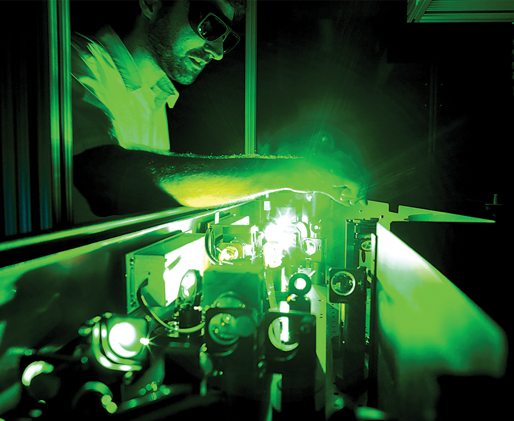 A photo shows a man wearing shades while operating an ultrafast optic laser in a dark room.