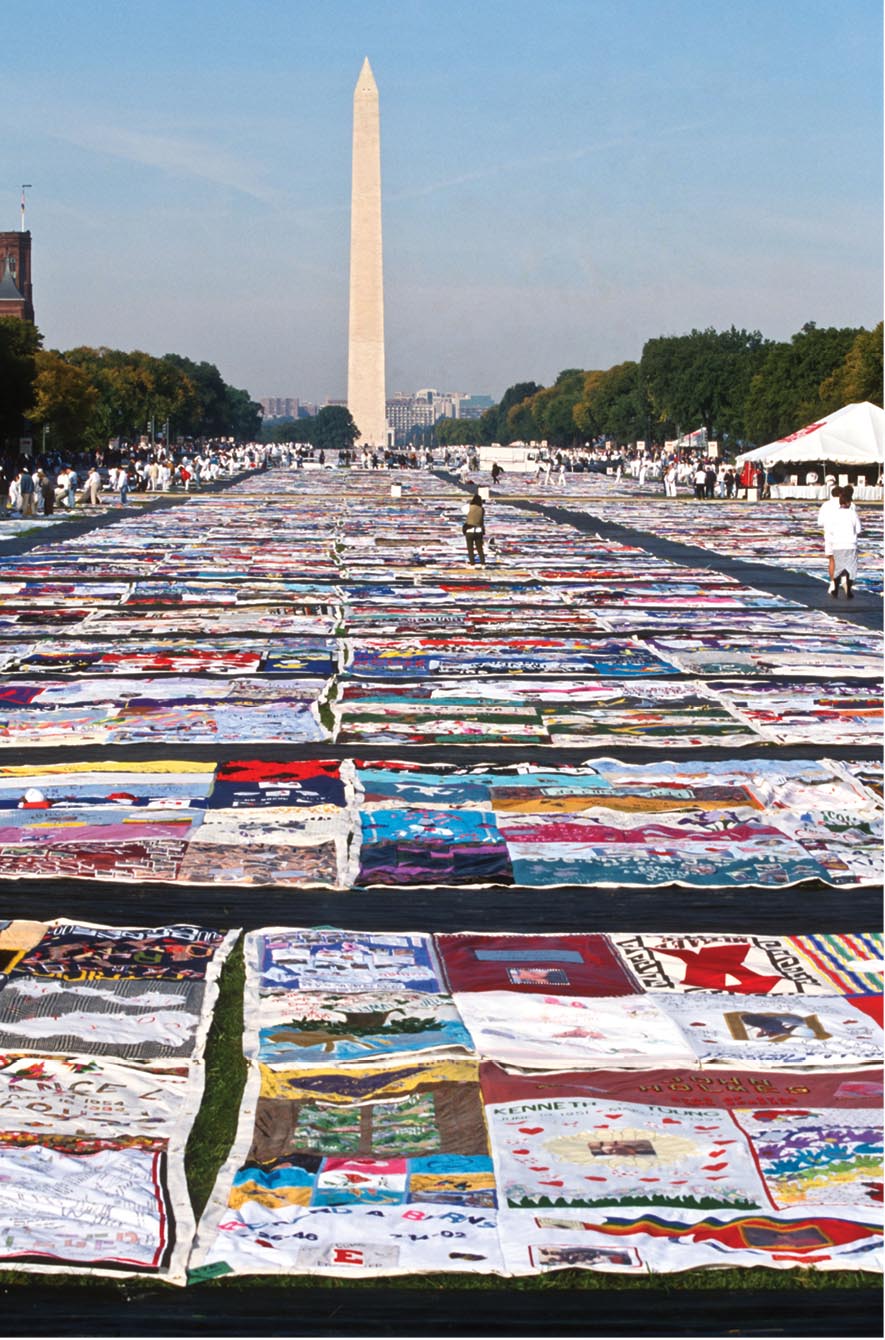 A number of quilts are spread out on the ground amongst a crowd in front of the Washington Monument.