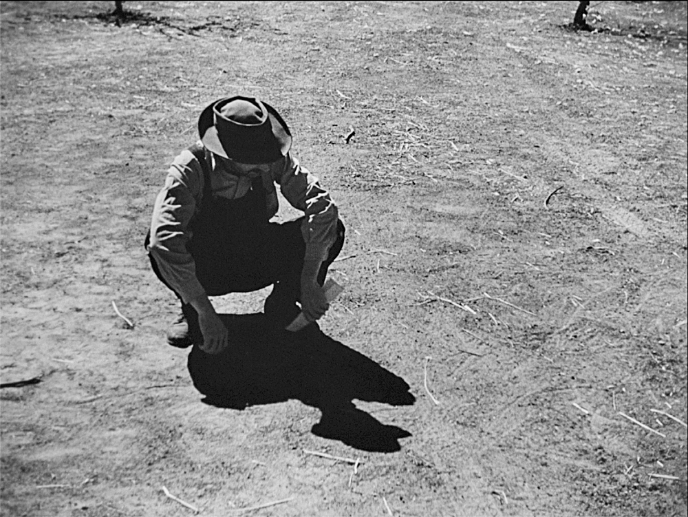 In The Grapes of Wrath, a man in a hat and overalls squats down to inspect a barren piece of land.