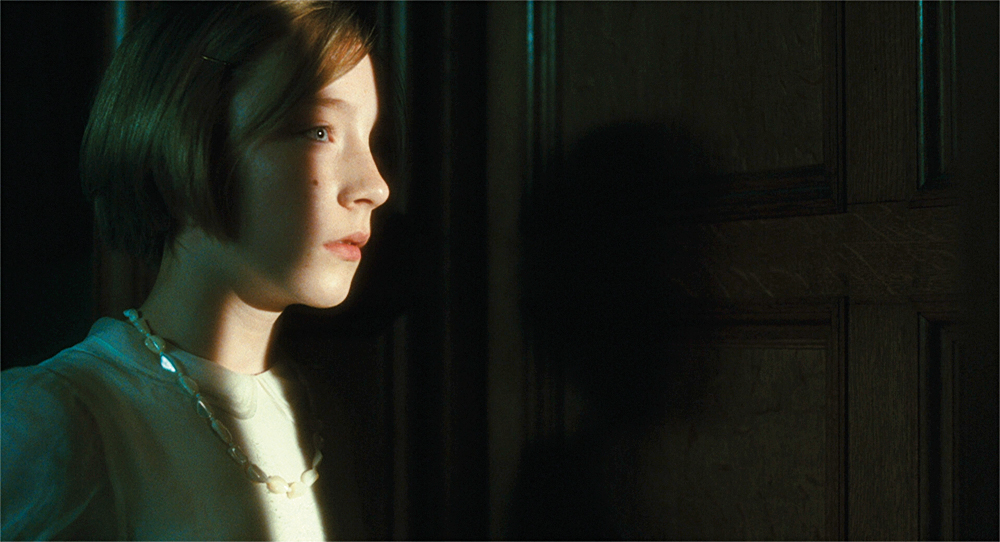A girl looks through a partially open door, and a thin strip of light illuminates her face in Atonement.