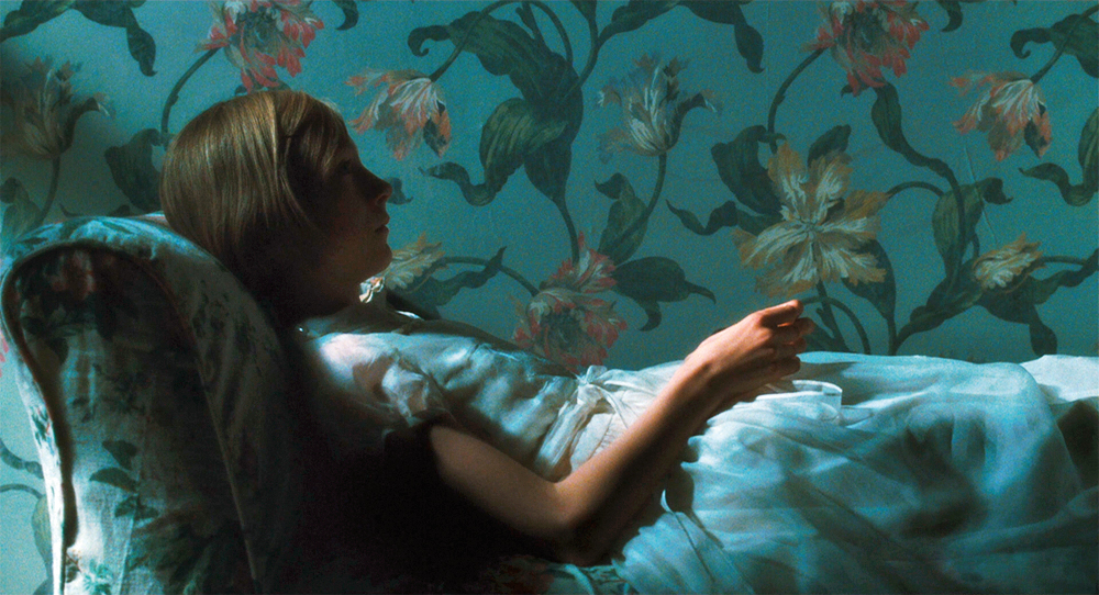 A girl in a dress lies on a couch in front of a wall which both have similar flowered patterns in Atonement.