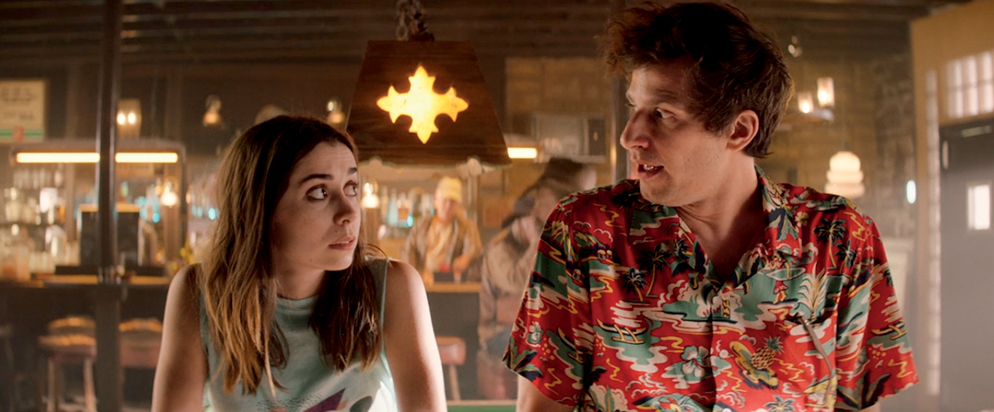 In Palm Springs, the camera shows a woman in a tank top and a man in a Hawaiian shirt mid-conversation at a bar.