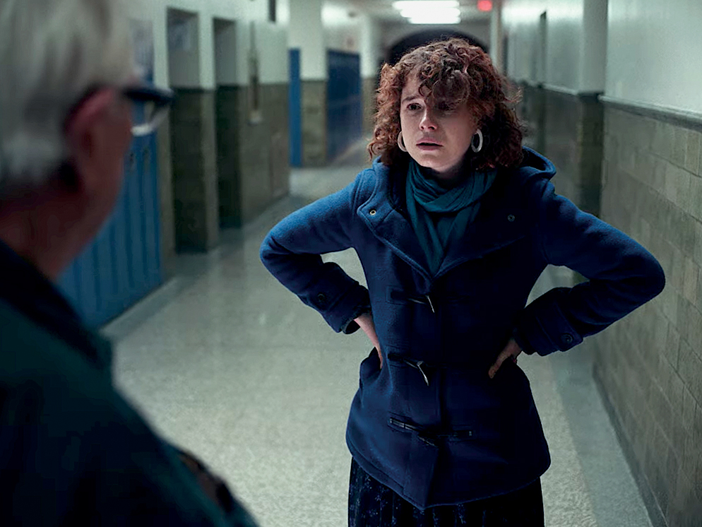 The protagonist of I’m Thinking of Ending Things stands in a hallway with her hands on her hips as she speaks to another character offscreen.
