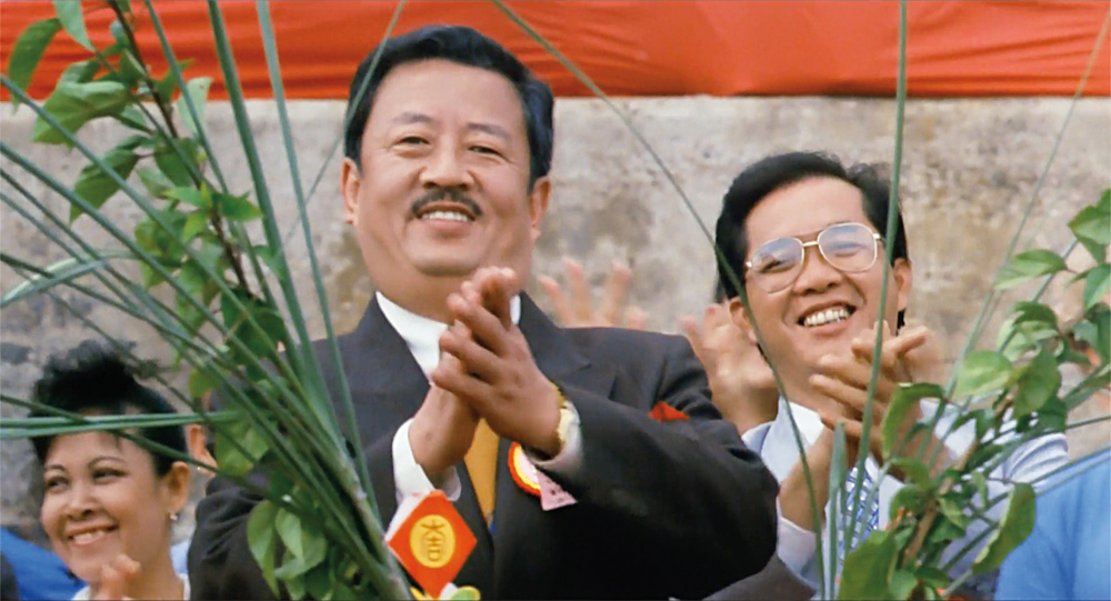 A man with a moustache in a suit and a group of people behind him smile and clap in The Killer.