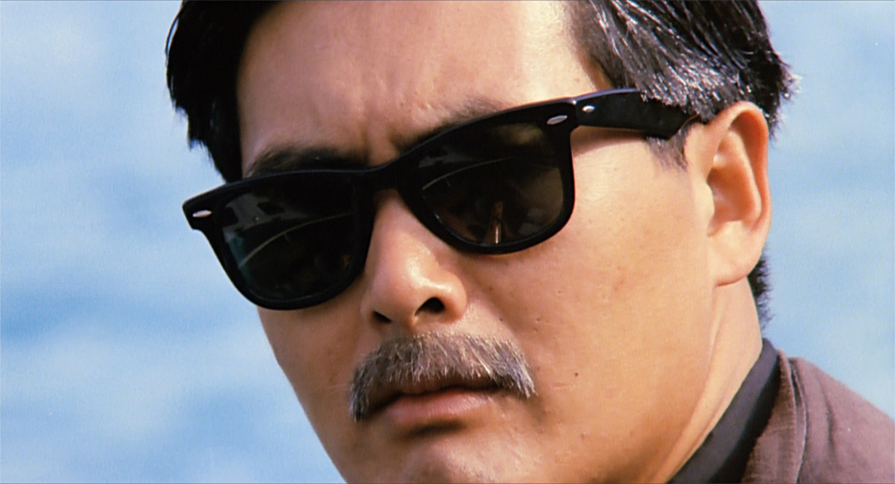 A man with sunglasses and a moustache pensively looks at the audience in The Killer.